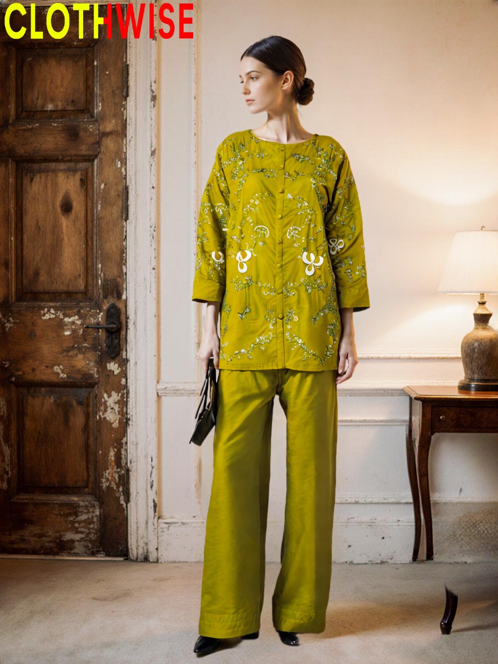 Woman wearing a green patterned outfit standing in a room with a wooden door and lamp.