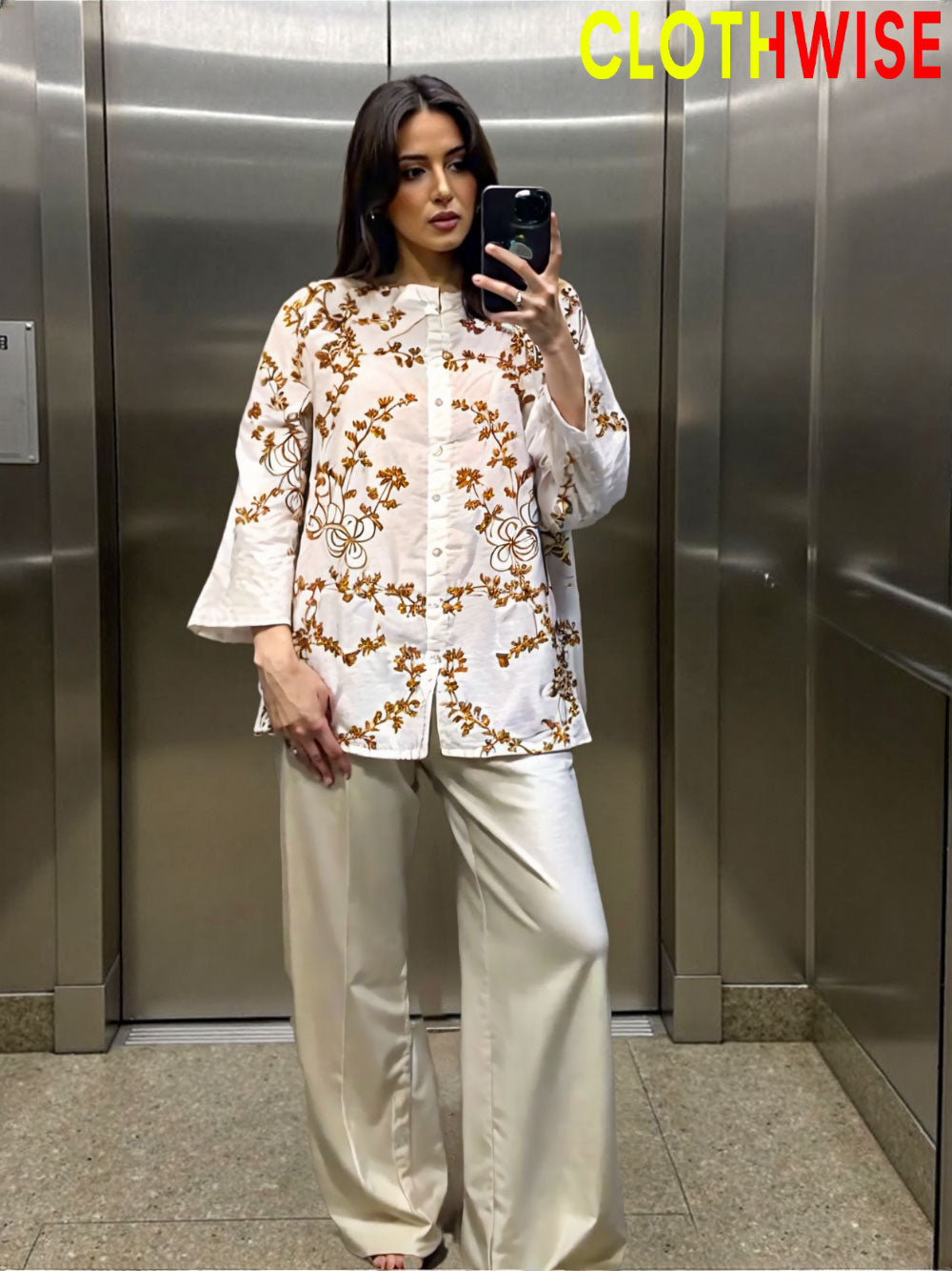 Woman taking a mirror selfie wearing a white floral blouse and beige pants in an elevator.