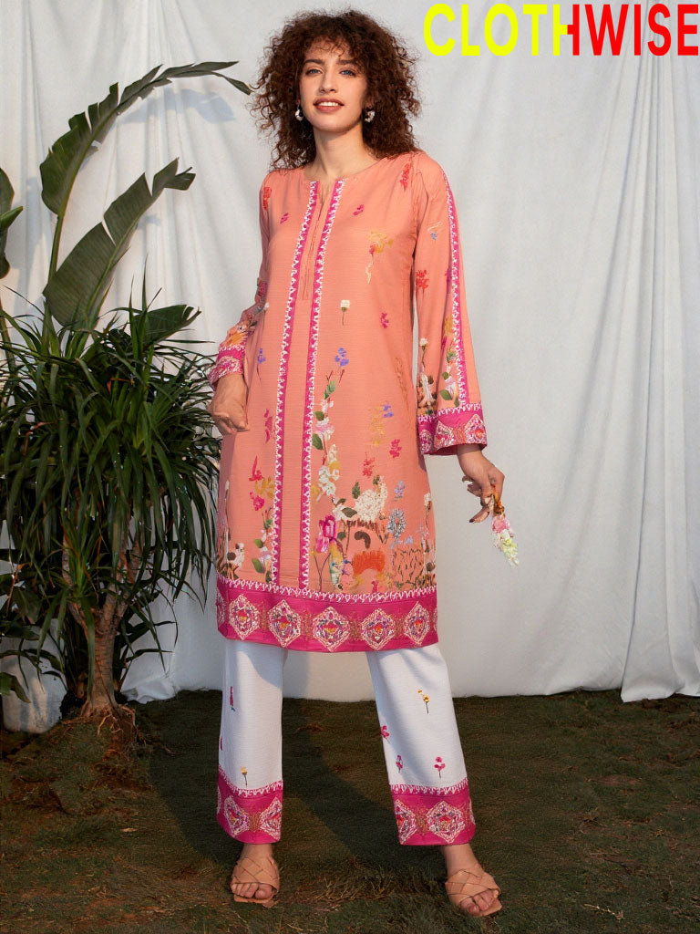 Woman wearing a peach embroidered top and white pants with pink borders, standing against a white curtain.