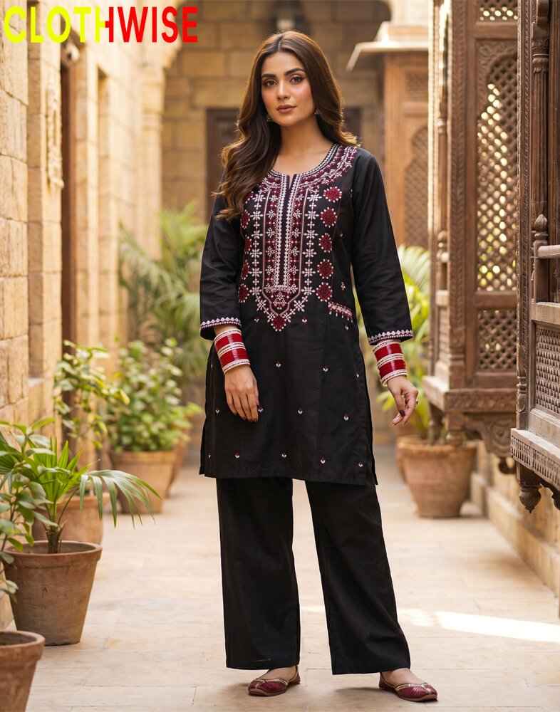 Woman wearing a black embroidered outfit in an outdoor setting with plants and architectural elements.