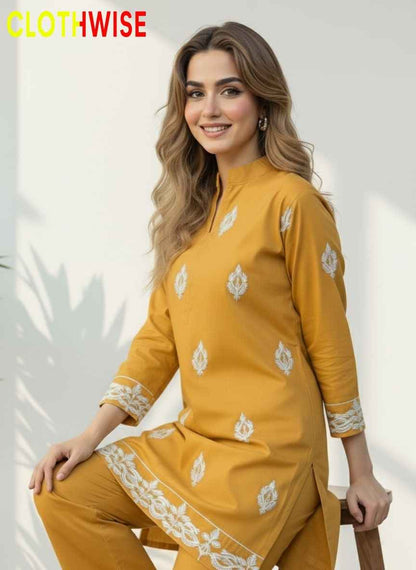 Woman wearing a mustard yellow traditional outfit with white patterns, sitting on a stool against a light background.