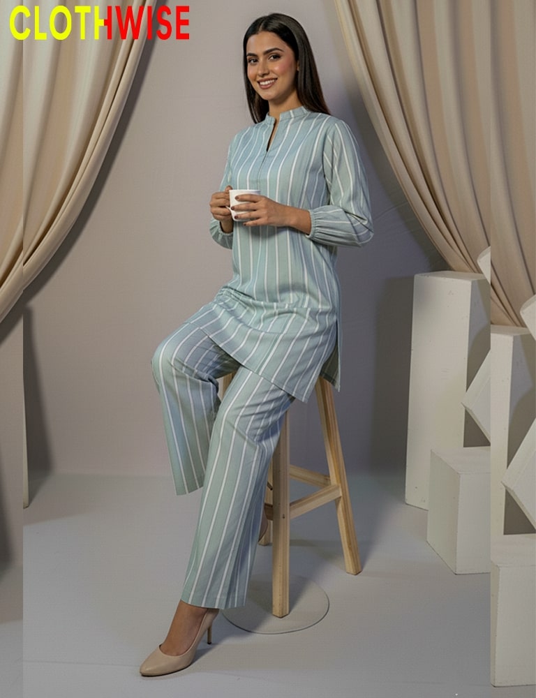 Woman in striped pajama set holding a cup, sitting on a stool with beige curtains in the background.