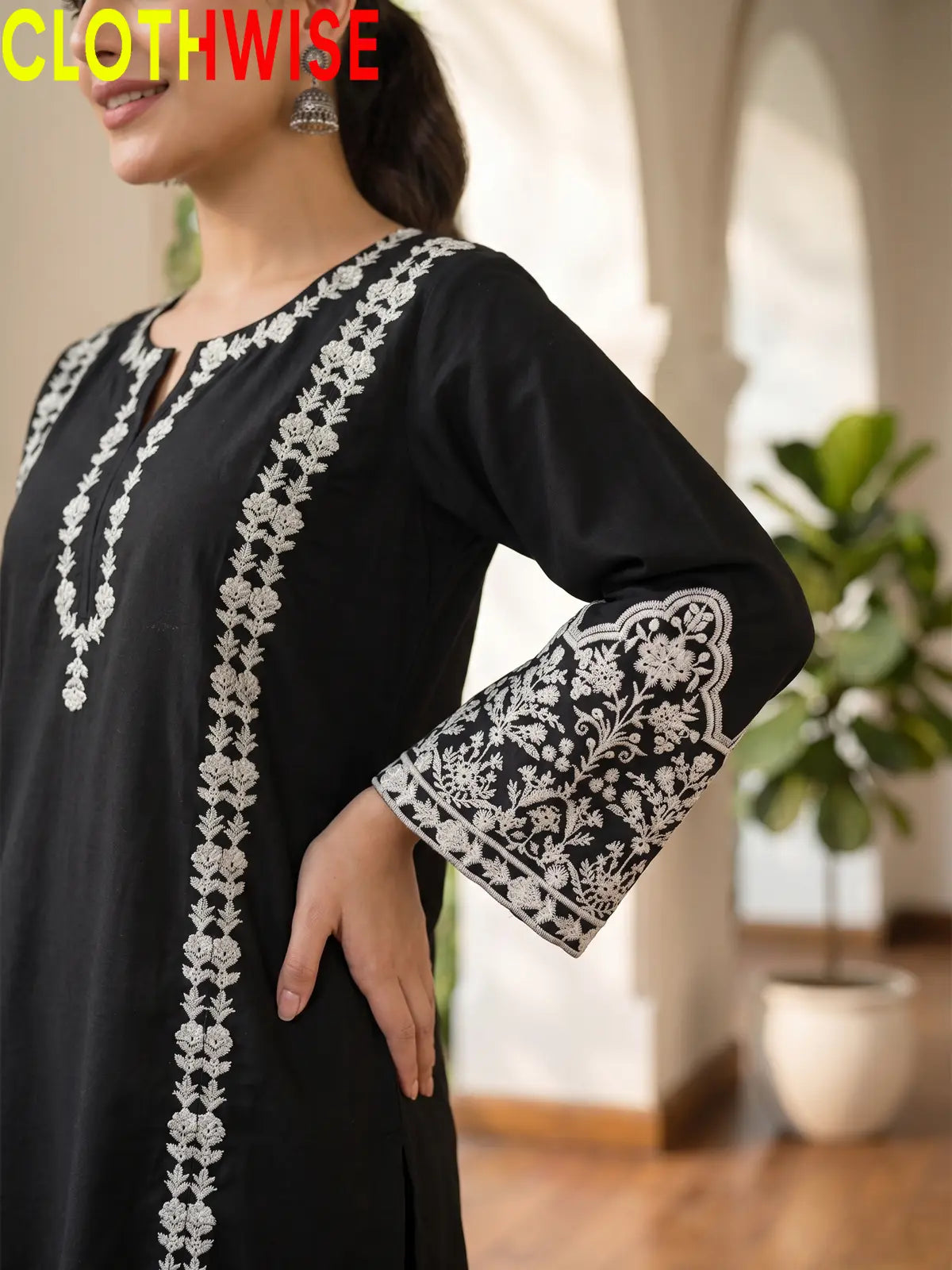 A woman wearing a black long kurta with intricate white floral embroidery on the neckline, front panel, and sleeve cuffs, shown in a softly lit indoor setting. The word CLOTHWISE appears in red at the top left.