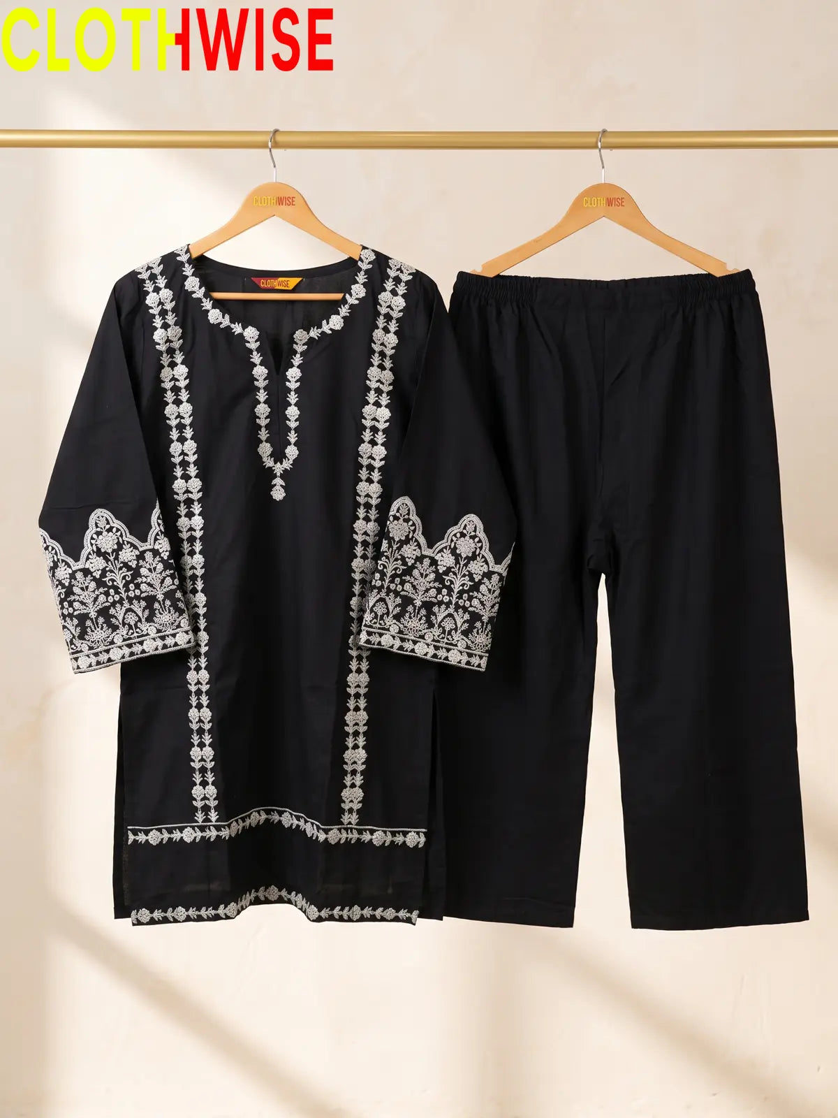 A black two-piece women's outfit set hanging on wooden hangers against a light beige wall. The set includes a long-sleeve tunic top with white floral embroidery on the sleeves, neckline, and hem, and matching black cropped pants.