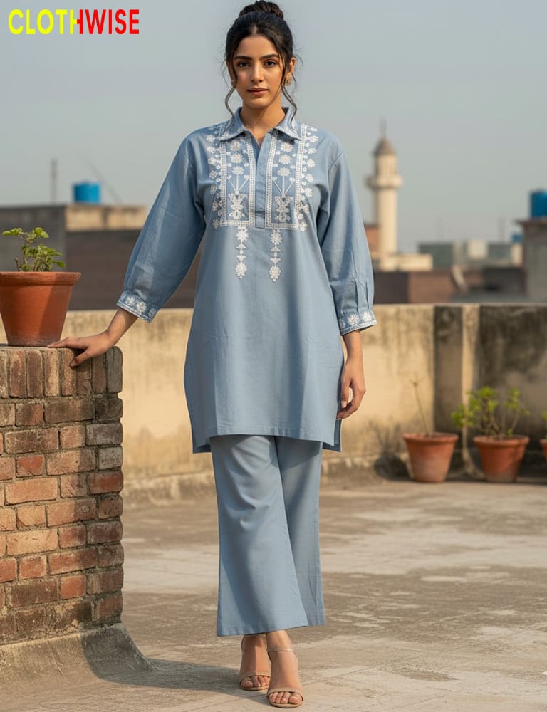 Woman wearing a light blue embroidered outfit on a rooftop with plants and a cityscape in the background.