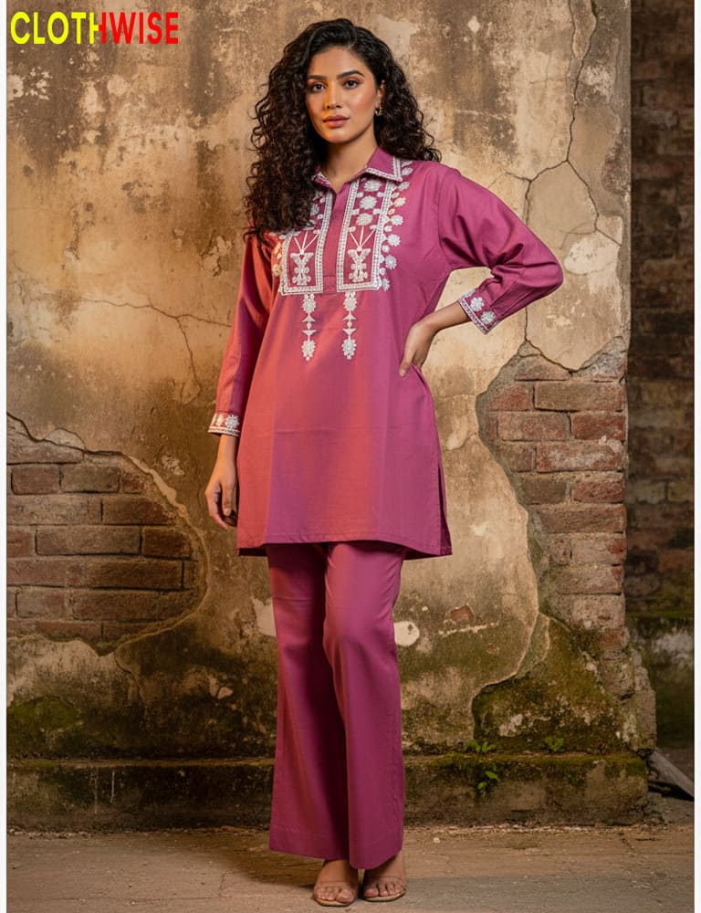 Woman wearing a pink traditional outfit with intricate designs against a textured wall.
