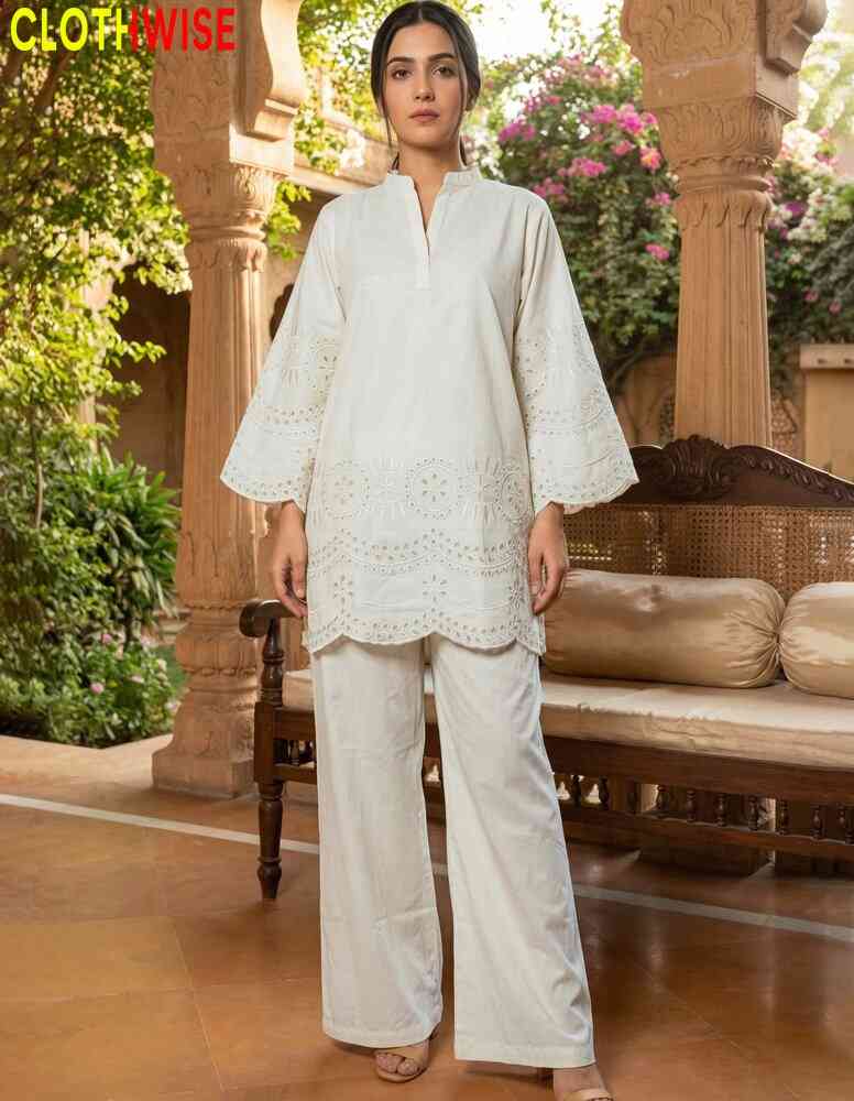 Woman wearing a white outfit with intricate patterns in an outdoor setting with floral decorations.