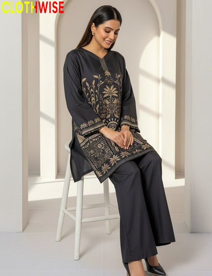 Woman wearing a dark gray embroidered outfit sitting on a white stool in a bright room.