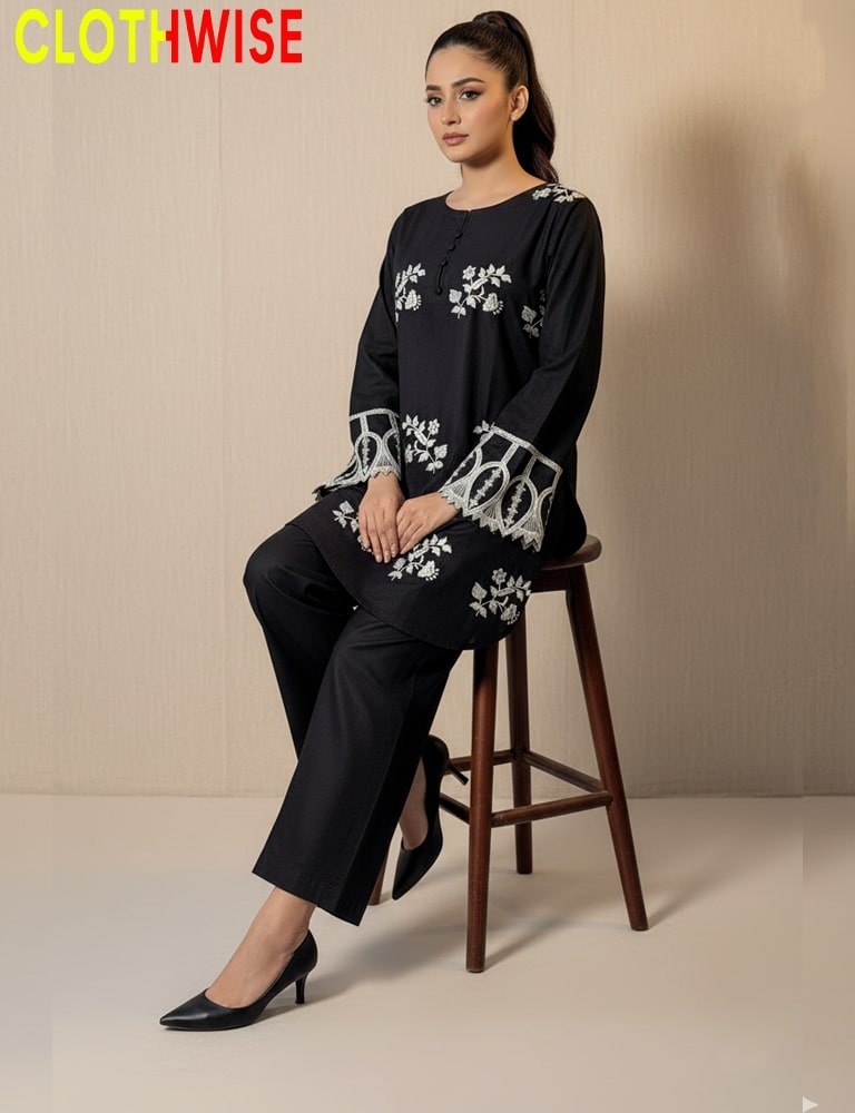 Woman wearing a black outfit with white floral embroidery sitting on a wooden stool against a beige background.