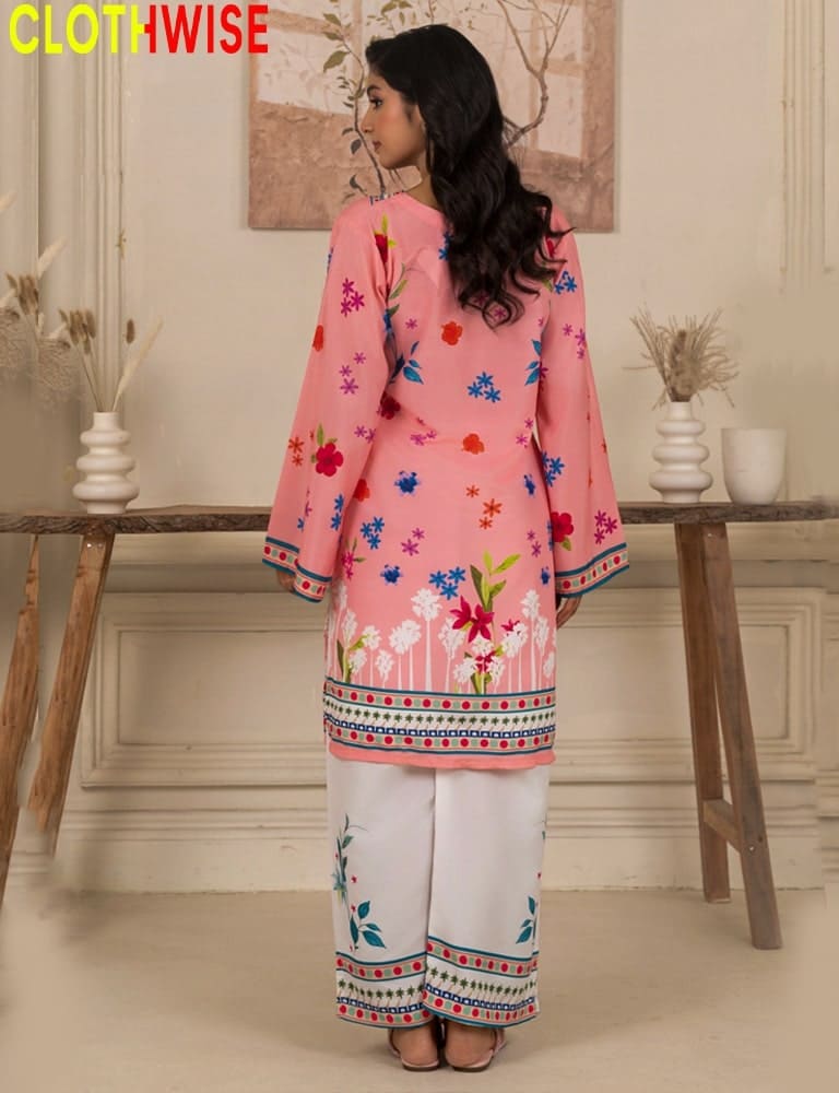 Woman wearing a pink floral top and white pants with decorative patterns, standing in a room with wooden furniture and decor.