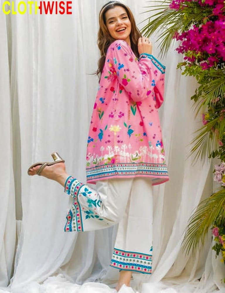 Woman wearing a pink floral outfit with white pants, standing against a white curtain with greenery.