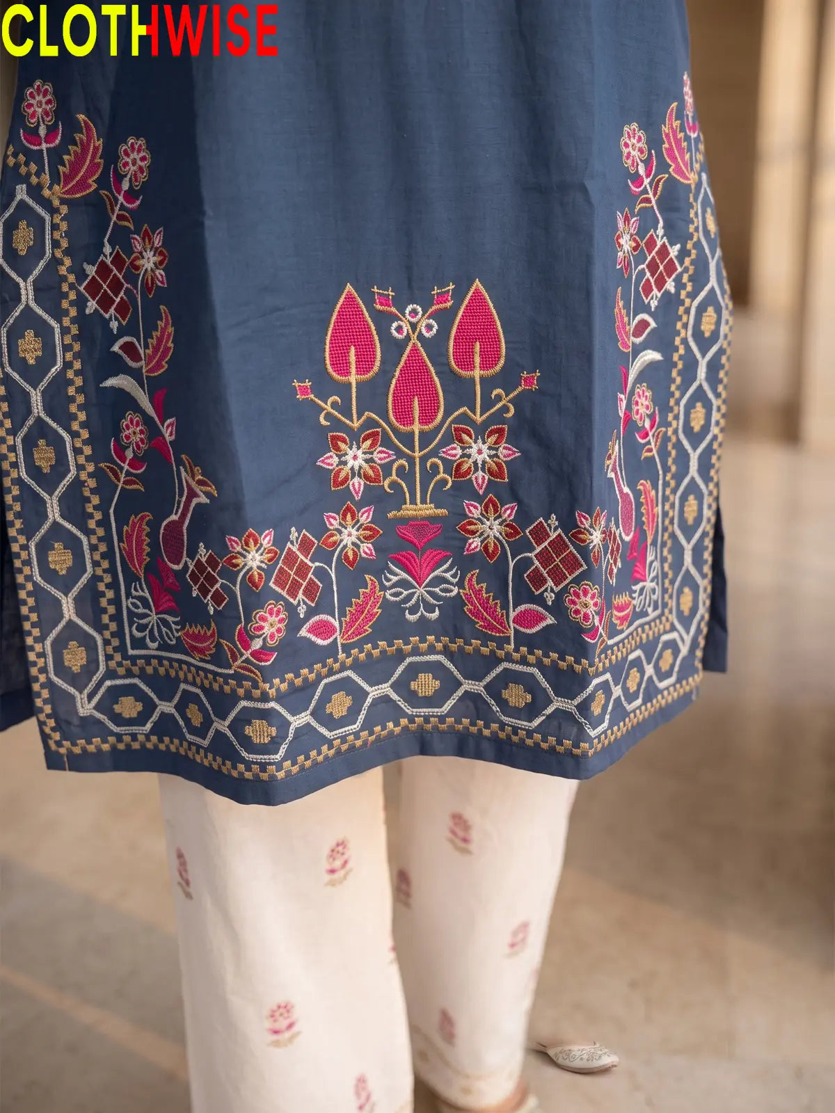 Close-up of a navy blue embroidered kurta fabric with pink, red, and gold floral and geometric border embroidery, paired with off-white printed pants. The brand name CLOTHWISE is visible in the top left corner.
