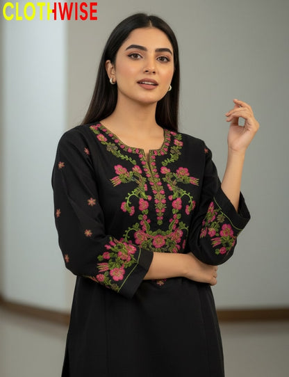 Woman wearing a black embroidered top with pink and green details, standing against a neutral background.