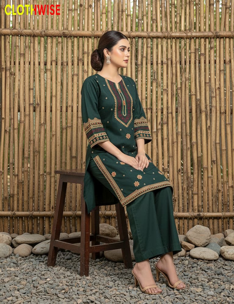 Woman in a green traditional outfit with intricate patterns sitting on a wooden stool against a bamboo wall.