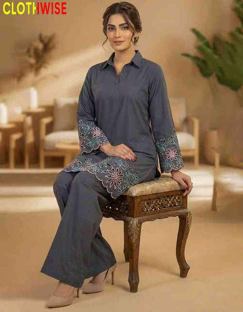 Woman in a gray traditional outfit with floral embroidery sitting on a wooden stool indoors.