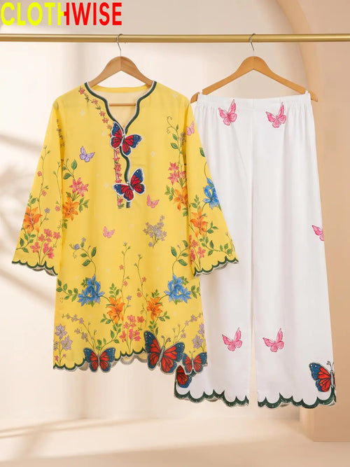 A two-piece women's pajama set hanging on wooden hangers against a warm beige background. The set includes a yellow short-sleeve tunic top with colorful butterfly and floral embroidery, and white pants with pink butterfly embroidery.