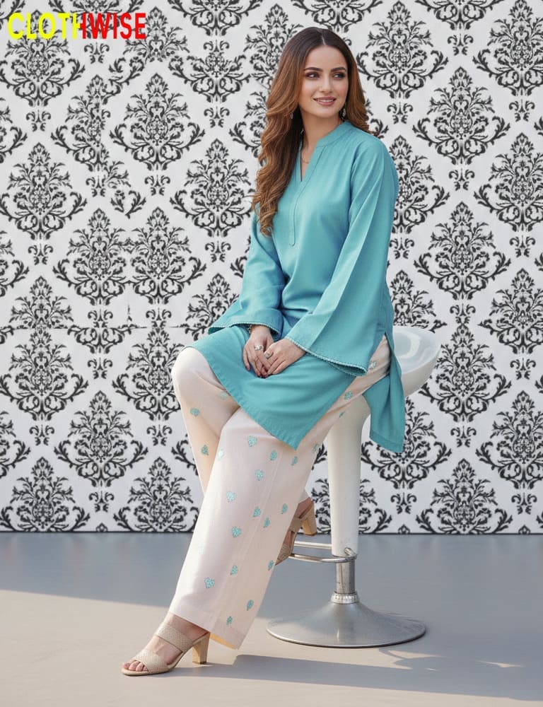 Woman in a blue kurta and white pants sitting on a stool against a patterned wall.