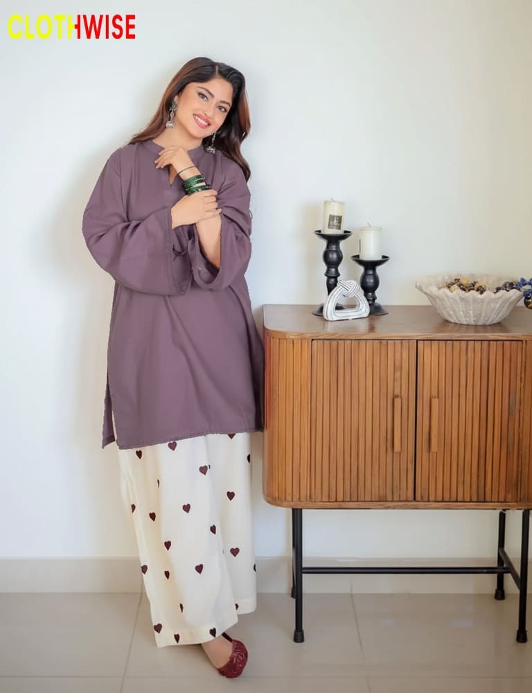 Woman in a purple top and white pants with heart patterns, holding a bottle, standing next to a wooden cabinet.