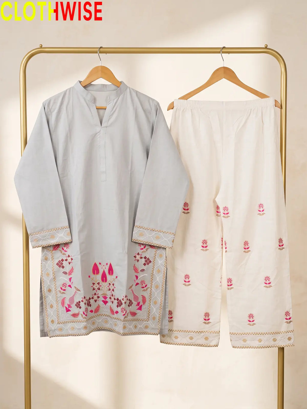 A two-piece women's outfit set hanging on a gold clothing rack. The set includes a light gray long-sleeve tunic with pink and gold floral embroidery at the hem, and matching off-white wide-leg pants with pink floral prints and gold borders.