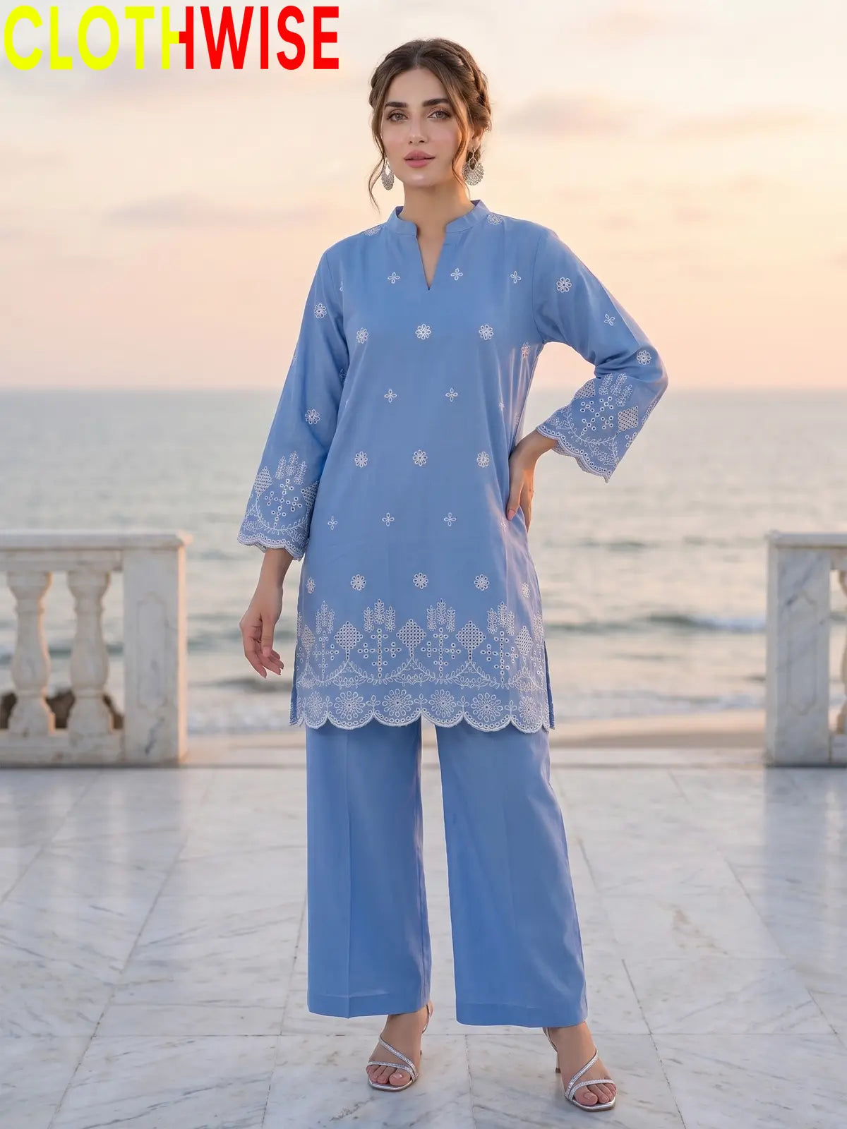 A woman modeling a light blue embroidered kurta and matching wide-leg pants set, standing on a marble walkway with the ocean and sunset in the background. The brand name 'CLOTHWISE' is displayed in the top left corner.