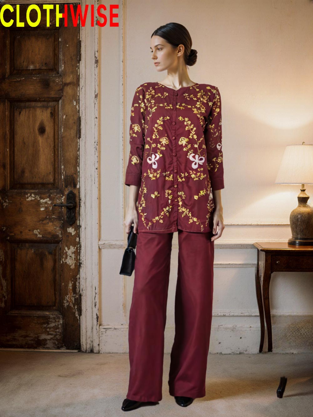 Woman wearing a maroon outfit with gold patterns in a room with wooden door and lamp.