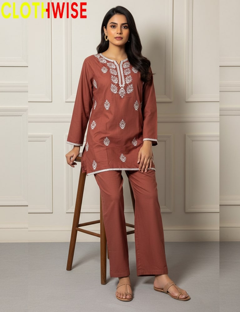 Woman wearing a rust-colored embroidered outfit on a wooden stool with a white paneled wall background.