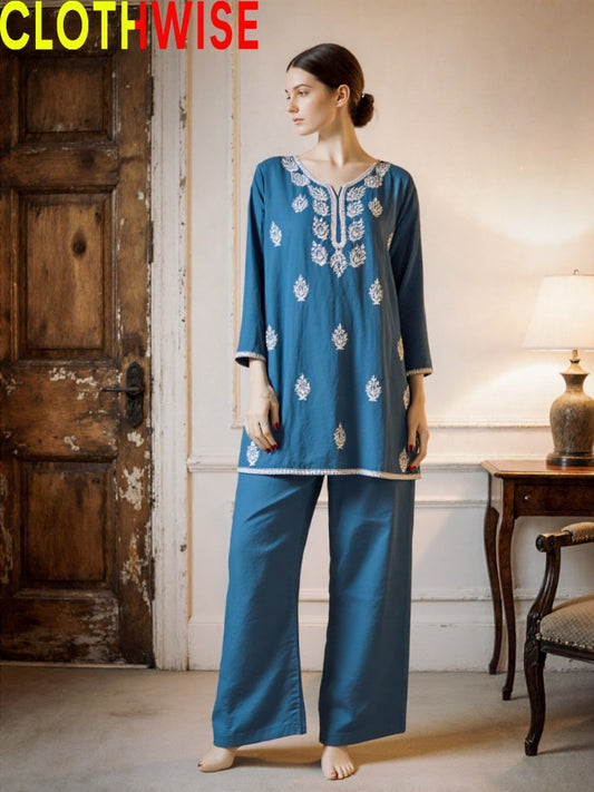 Woman wearing a blue embroidered outfit in a room with wooden door and lamp.