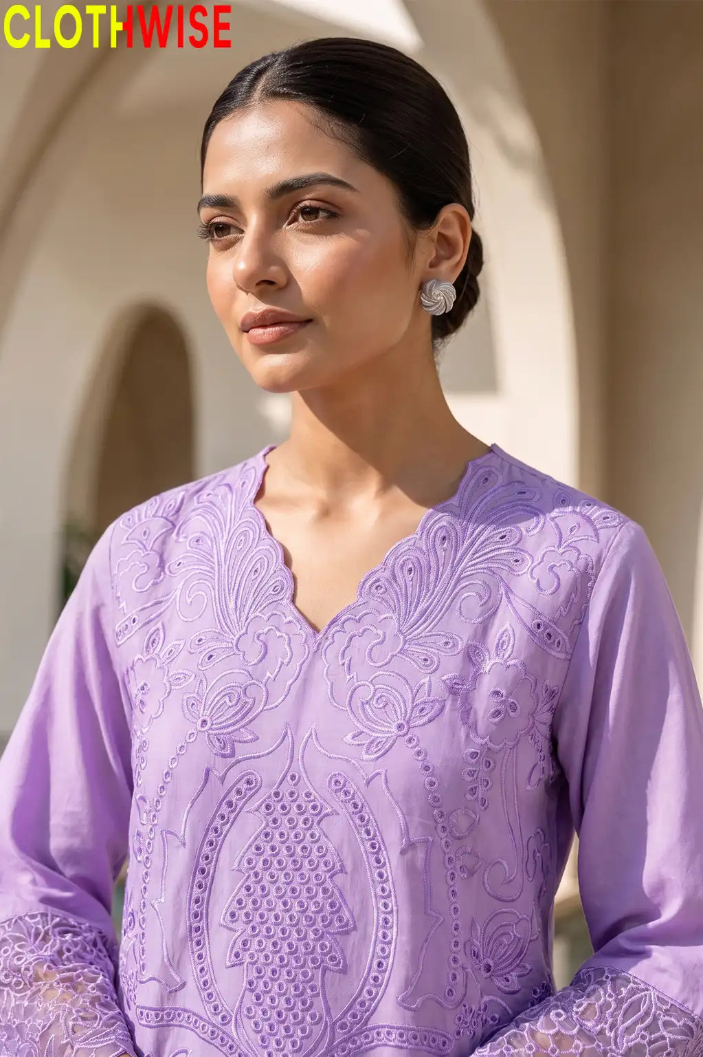 A woman wearing a lavender embroidered kurta with a scalloped V-neckline and intricate floral embroidery, shown in a warm-toned architectural setting. The CLOTHWISE logo is visible in the top left corner.