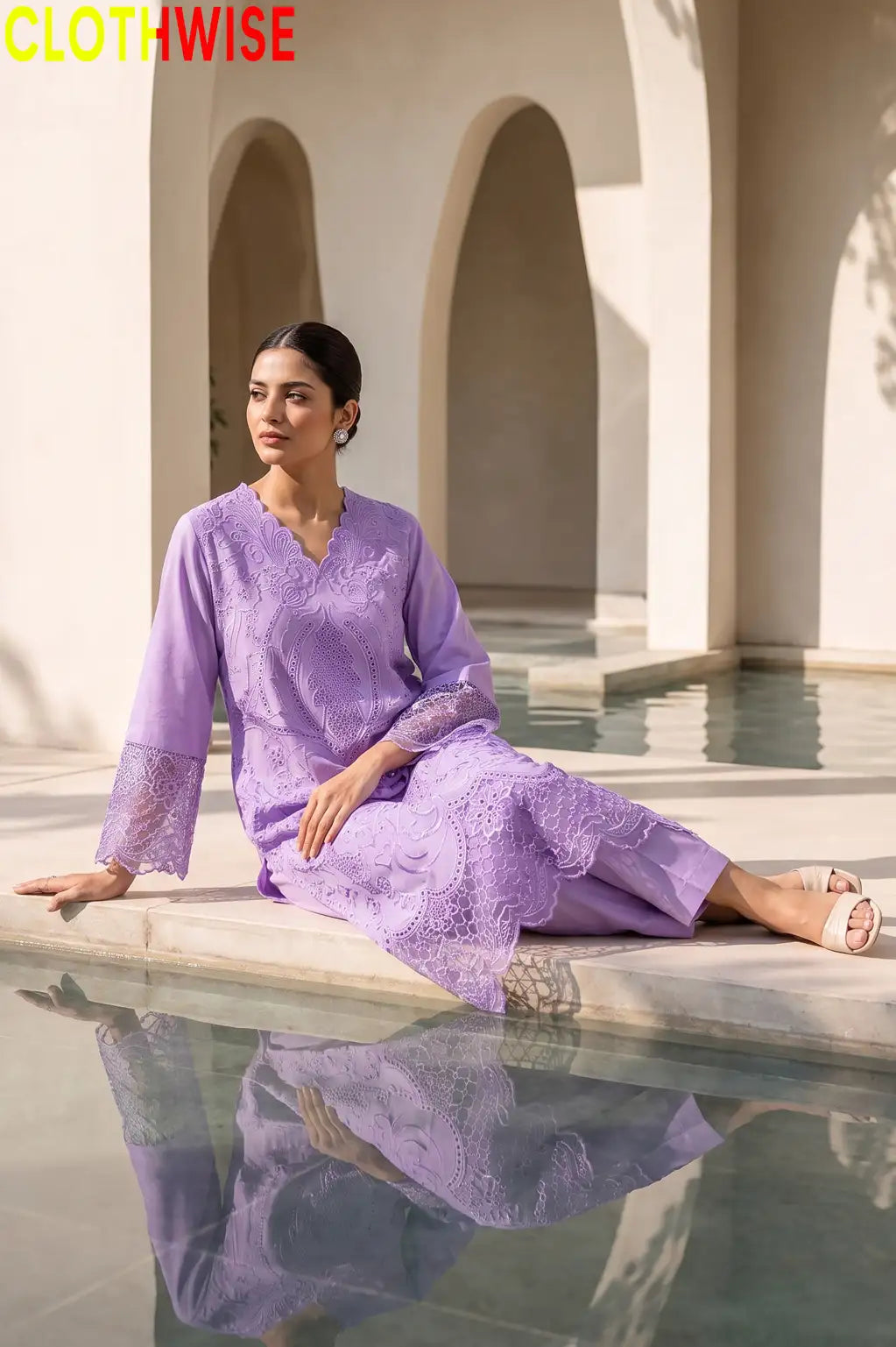 A woman wearing a lavender purple embroidered kurta set sits on the edge of a reflecting pool in a sunlit architectural setting with arches. The brand name CLOTHWISE is displayed in the top left corner.