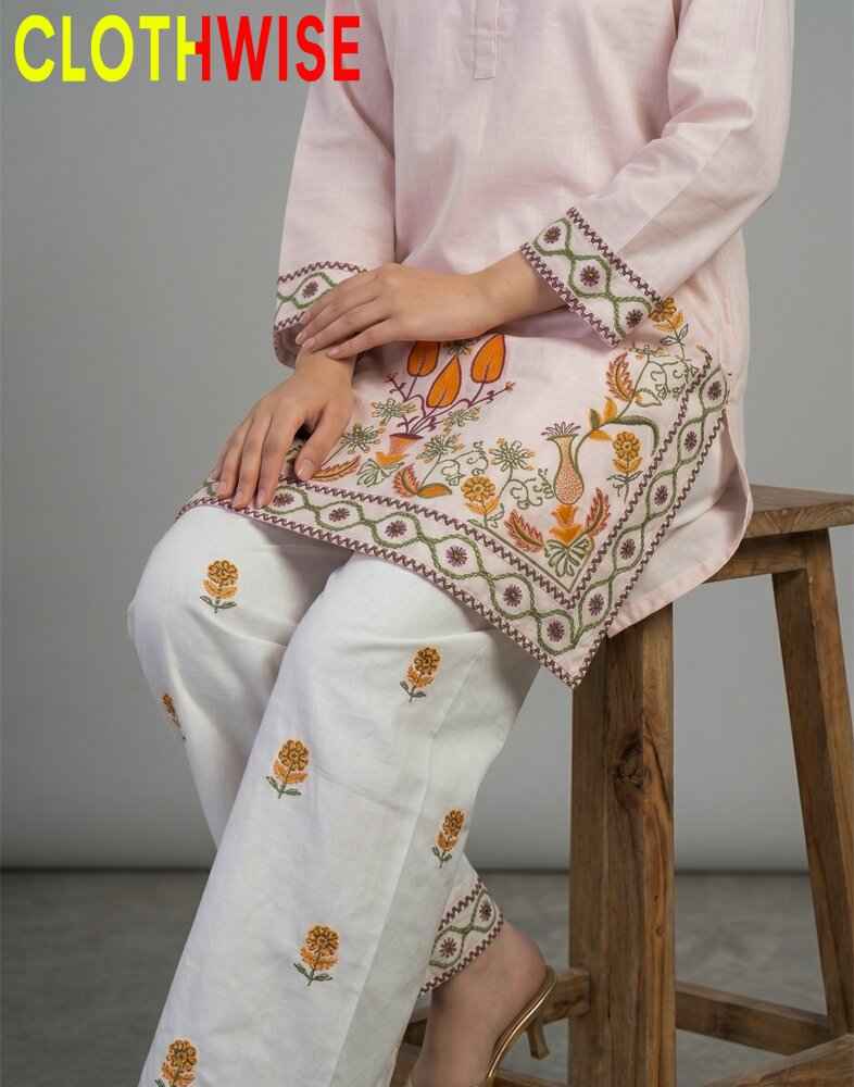 Person wearing a light pink kurta with white pants featuring intricate embroidery, sitting on a wooden stool.