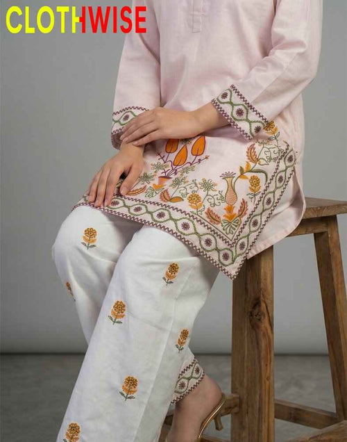 Person wearing a light pink kurta with white pants featuring intricate embroidery, sitting on a wooden stool.