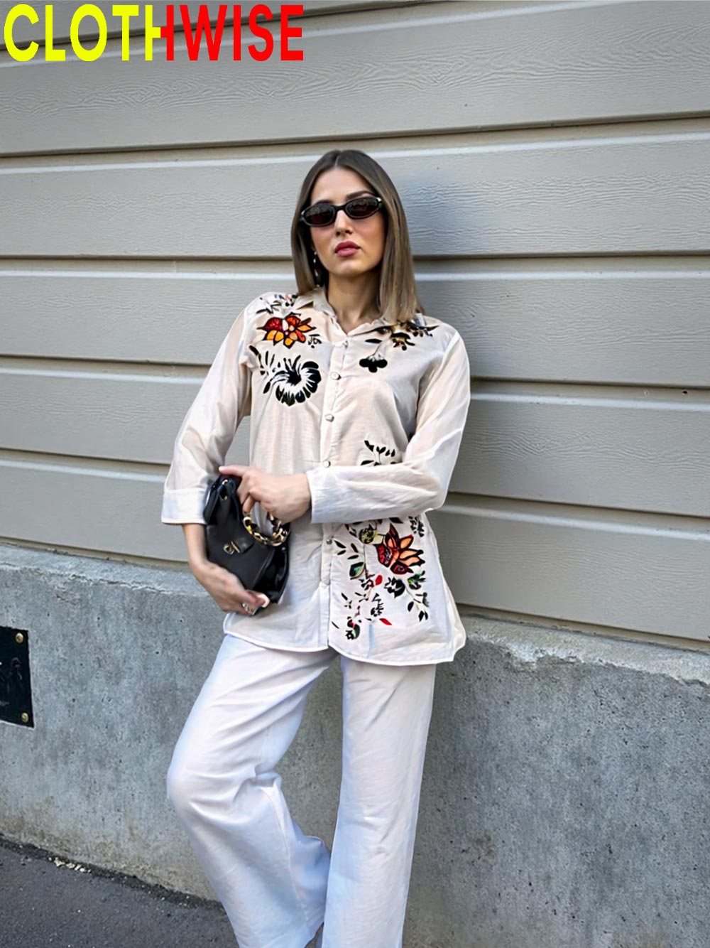 Woman wearing a stylish outfit with an embroidered blouse and white pants against a gray wall.