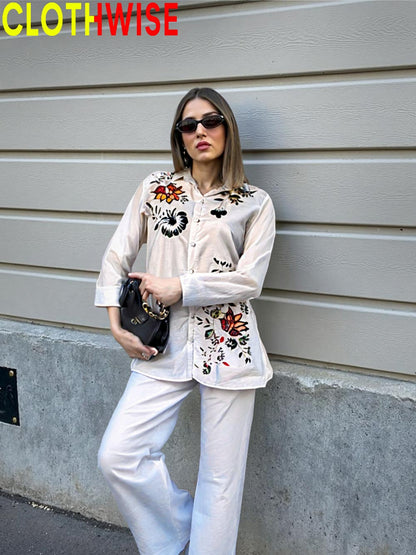 Woman wearing a stylish outfit with an embroidered blouse and white pants against a gray wall.