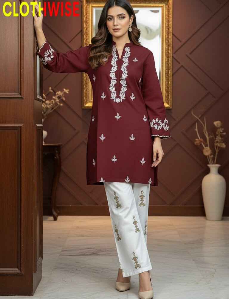 Woman wearing a maroon top and white pants with embroidery, standing in a room with wooden paneling and a vase.