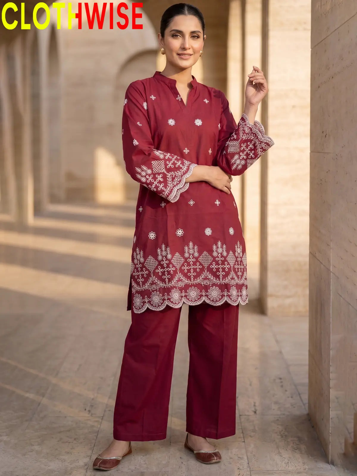 A woman modeling a maroon embroidered kurta and matching wide-leg pants set, standing on a marble walkway with the ocean and sunset in the background. The brand name 'CLOTHWISE' is displayed in the top left corner.