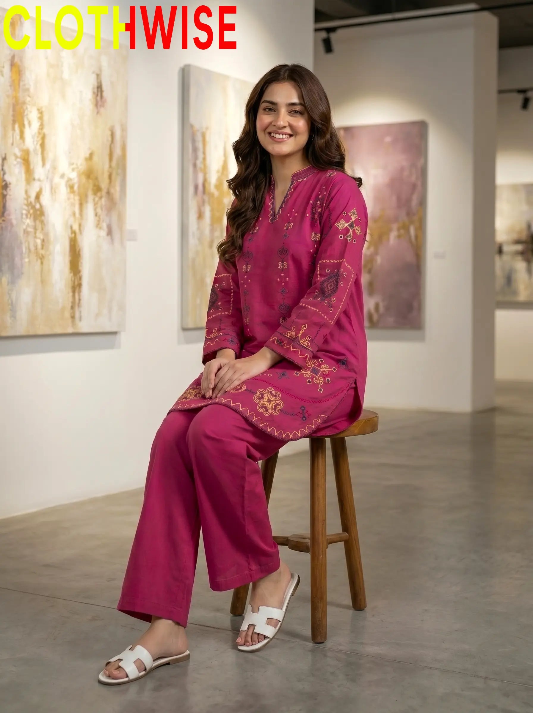 Woman in a pink embroidered outfit sitting on a stool in an art gallery with 'CLOTHWISE' branding.
