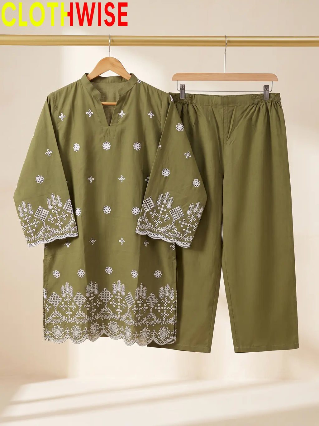 An olive green two-piece clothing set hanging on wooden hangers against a warm beige background. The set includes a long-sleeve tunic top with white embroidered floral and geometric patterns, and matching straight-leg pants.