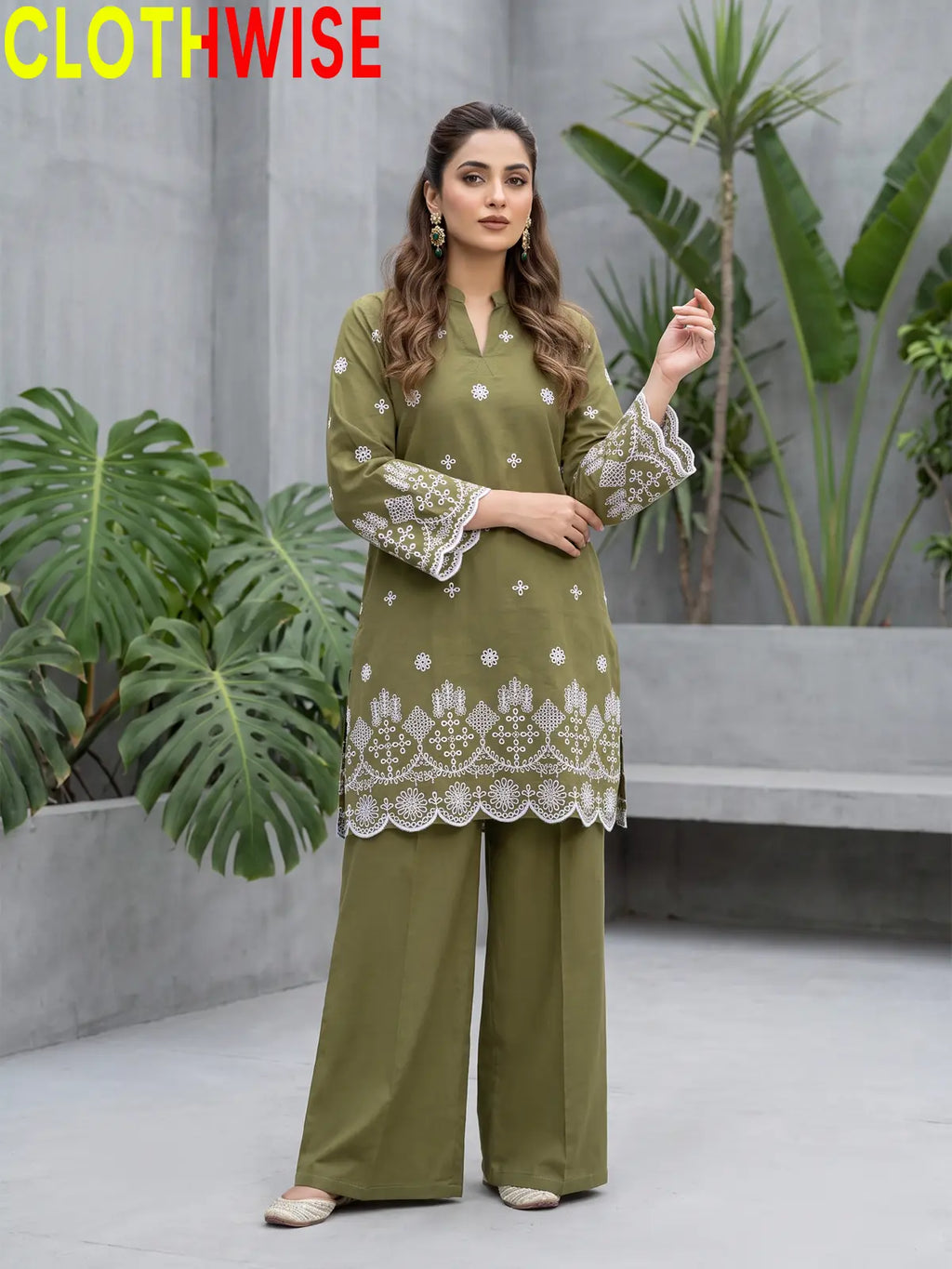 A woman modeling a moss green embroidered kurta and matching wide-leg pants set, standing on a marble walkway with the ocean and sunset in the background. The brand name 'CLOTHWISE' is displayed in the top left corner.