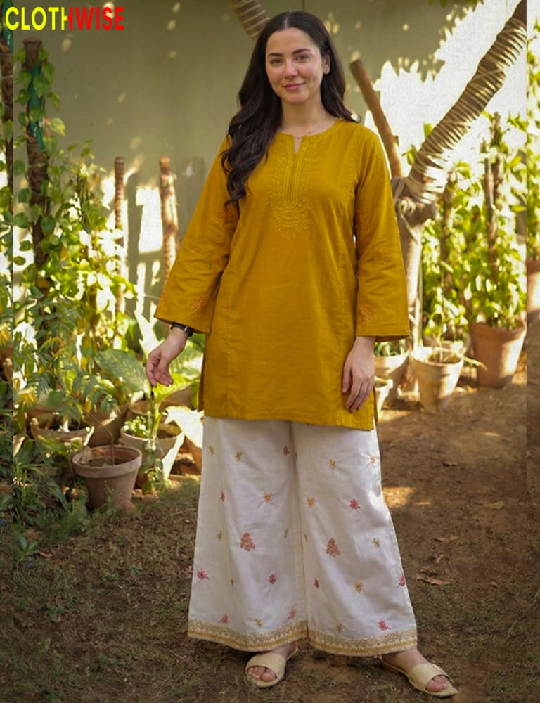 Woman wearing a mustard yellow top and white pants with floral patterns, standing in a garden.