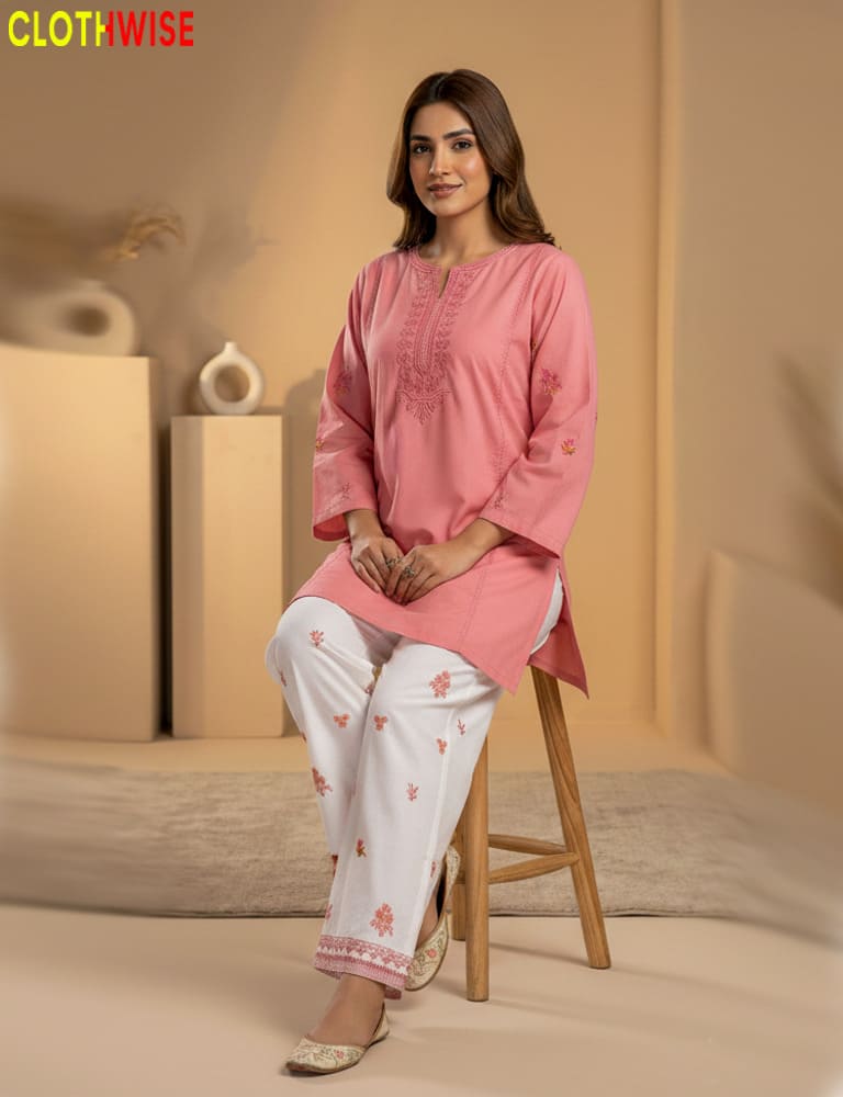 Woman wearing a pink kurta with white pants sitting on a stool against a beige background.