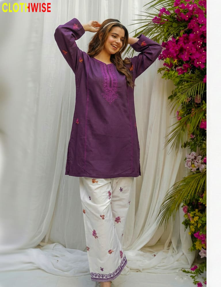 Woman wearing a purple embroidered top and white pants with floral patterns, standing against a white curtain with floral decorations.