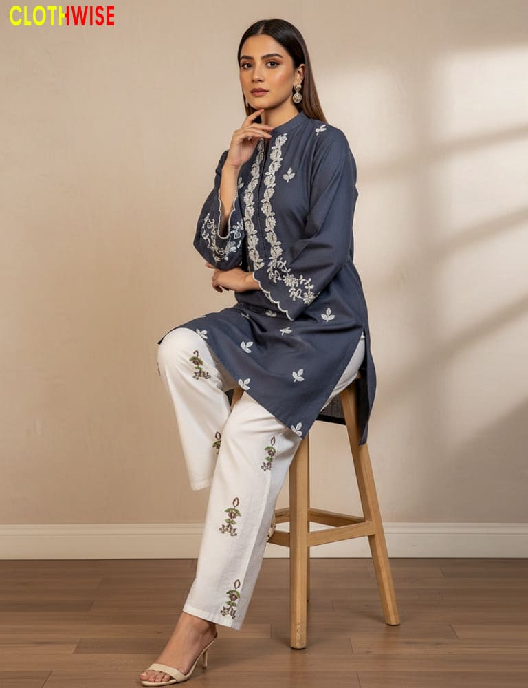 Woman wearing a navy blue embroidered top and white pants sitting on a wooden stool against a beige wall.
