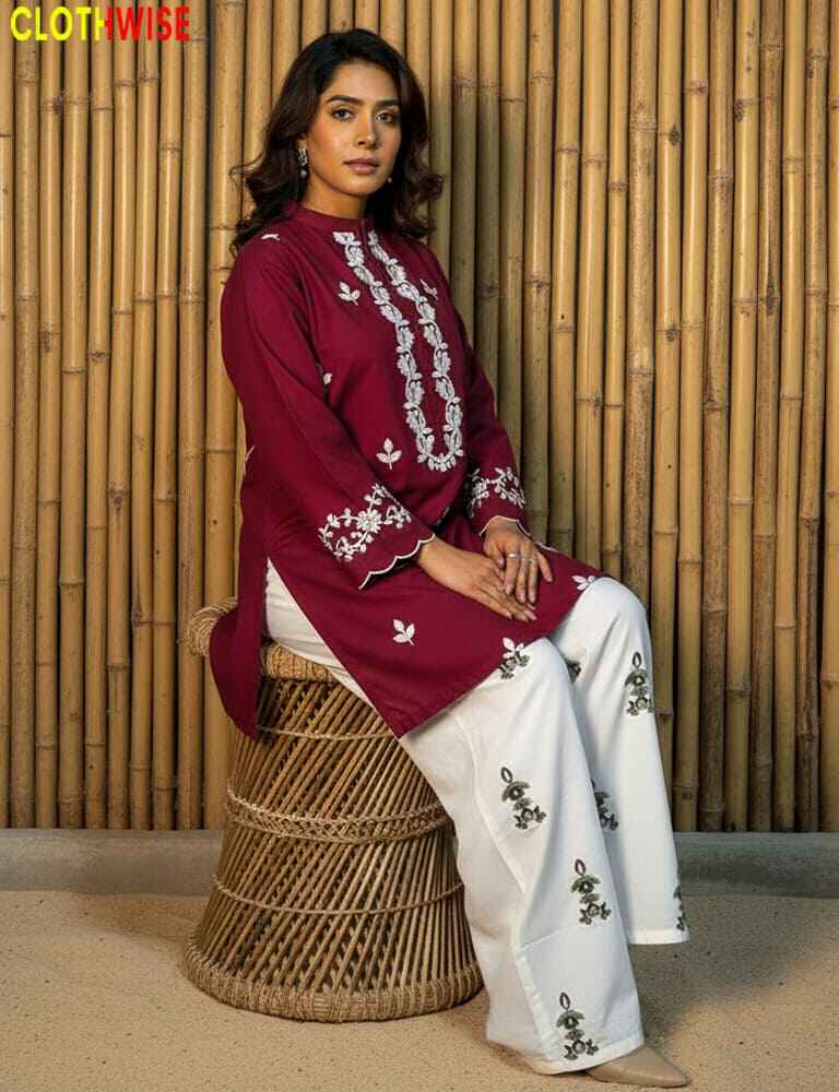 Woman wearing a maroon and white embroidered outfit sitting on a woven stool against a bamboo wall.