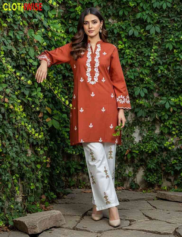 Woman wearing a rust-colored embroidered top and white pants standing against a green leafy background.