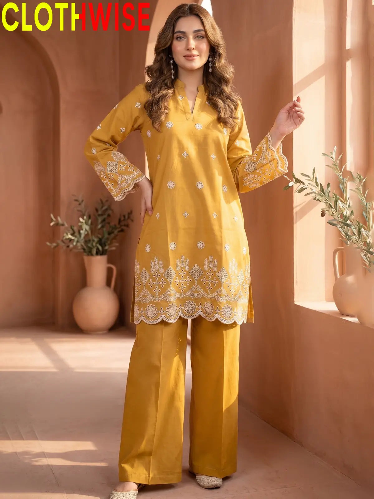A woman models a mustard yellow two-piece kurta and trouser set with white floral embroidery, set against a warm, sunlit desert architectural background. The brand name 'CLOTHWISE' is displayed in the top left corner.