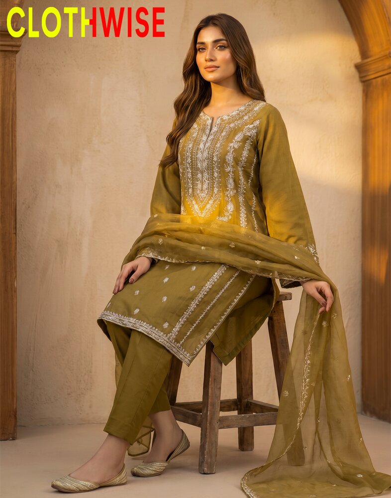 Woman wearing a green traditional outfit with embroidery, sitting on a wooden stool against a beige wall.