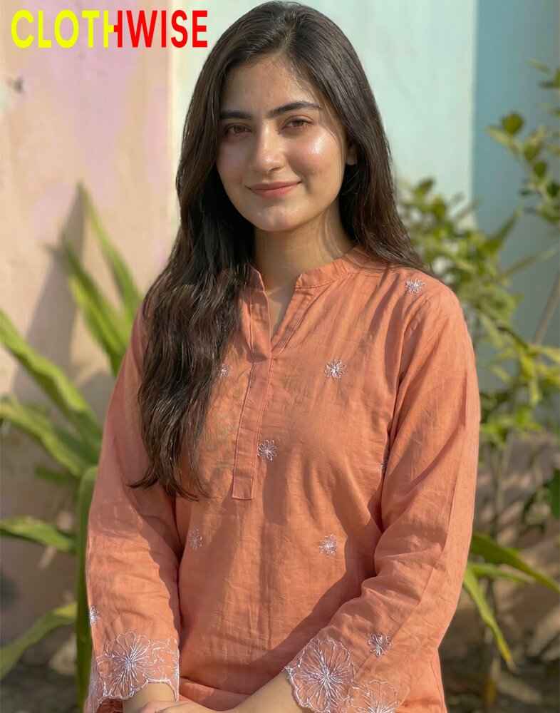 Woman wearing an orange kurta with floral patterns, standing outdoors with greenery in the background.