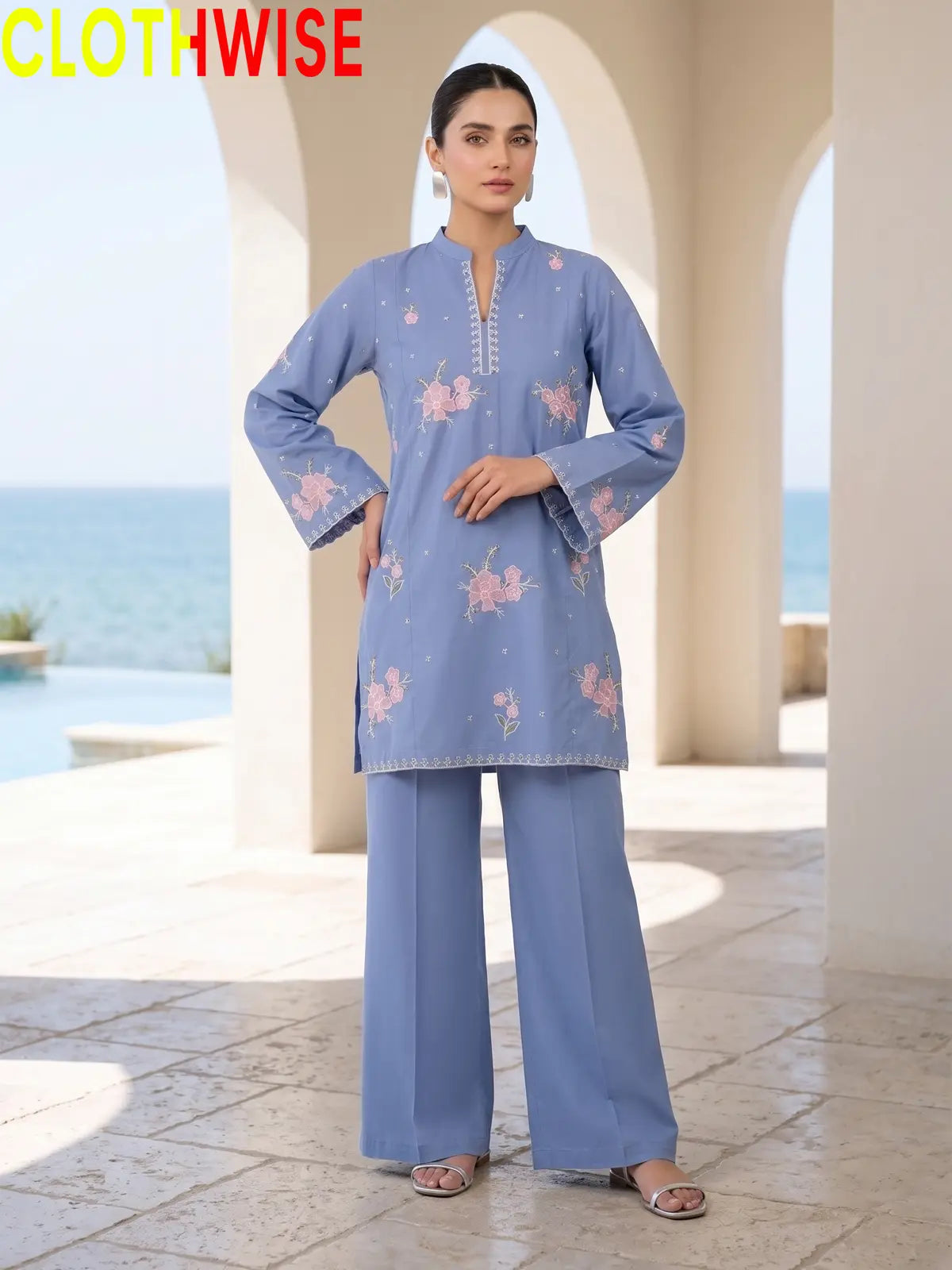 Front view of a model wearing a periwinkle blue embroidered straight tunic and matching palazzo pants against an outdoor arches backdrop