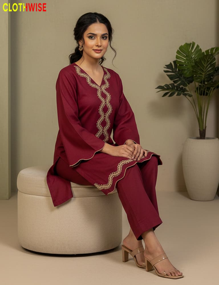 Woman in a maroon outfit with gold embroidery sitting on a beige stool against a neutral background.