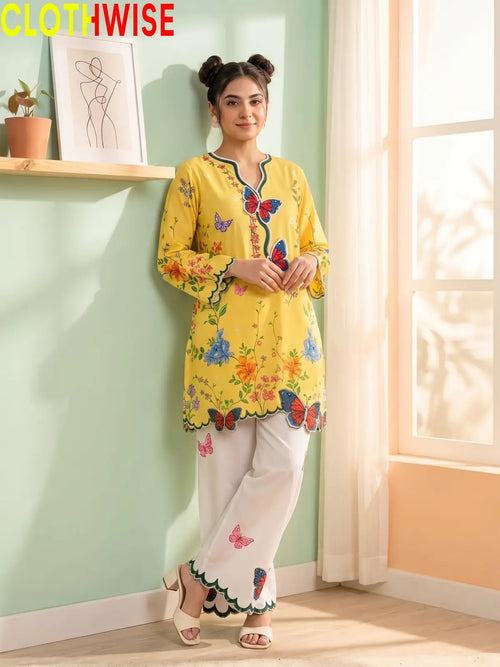 A woman models a yellow embroidered kurta with colorful butterfly and floral motifs, paired with white wide-leg pants, in a brightly lit room with a mint green wall and window.